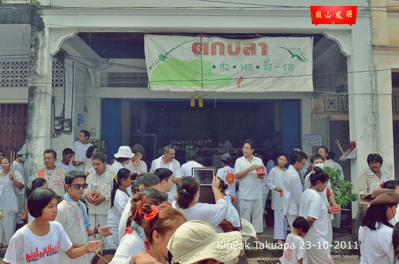 ชาวบ้านชุดขาวรวมตัวหน้าร้านตึกแถวระหว่างพิธีบนถนน