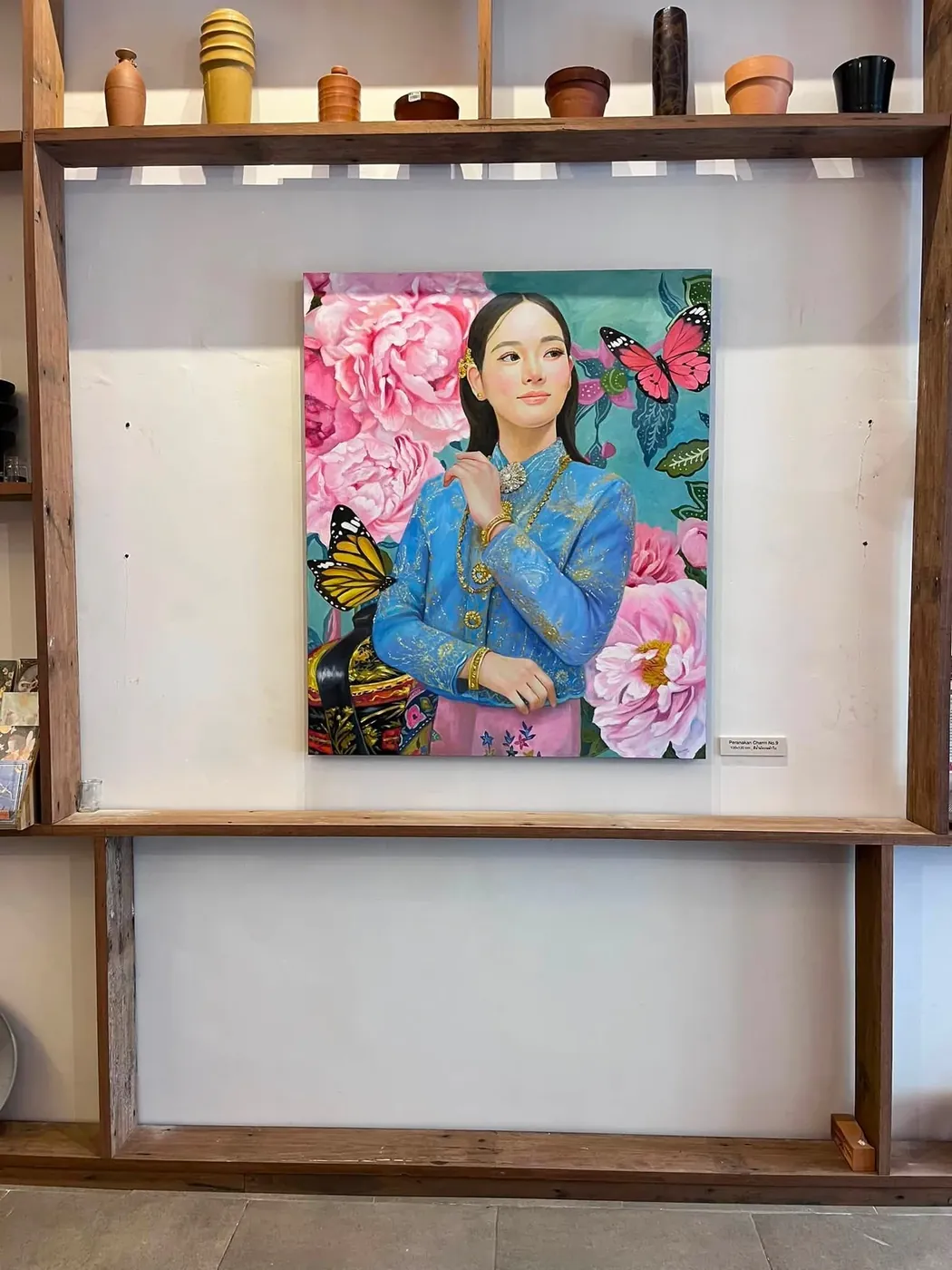ภาพเขียน Peranakan Charm No.9 แขวนอยู่บนชั้นไม้ในนิทรรศการ พร้อมป้ายกำกับขนาดผลงาน